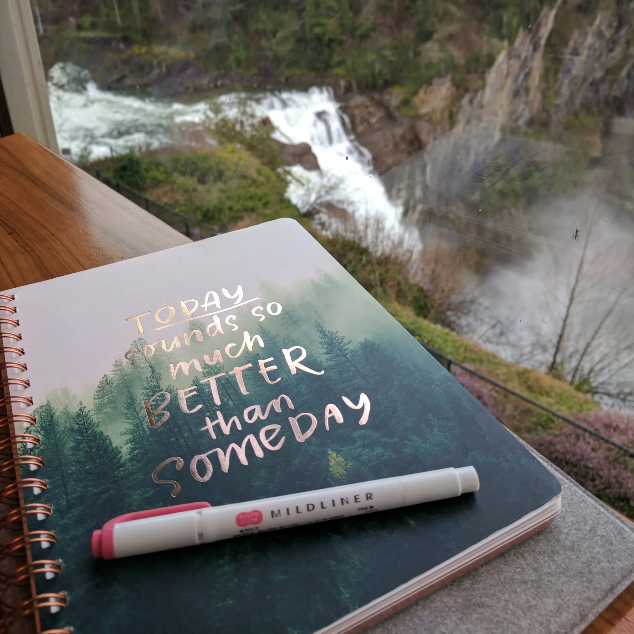 A journal and highlighter on a table near a window overlooking a waterfall.