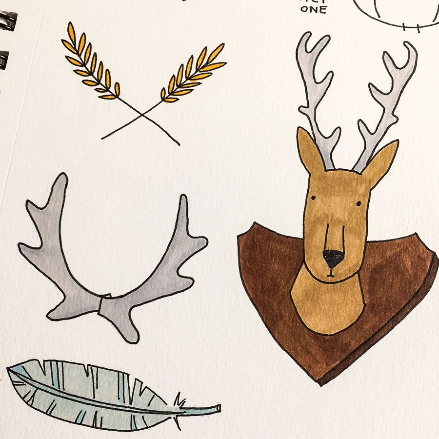 A cartoon style deer with antlers pictured on a bullet journal practice page next to a sprig of wheat and a hand-drawn feather.