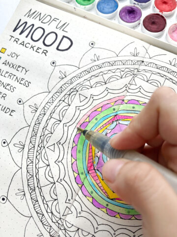 A hand coloring a circle on a bullet journal mandala during mindfulness practice.
